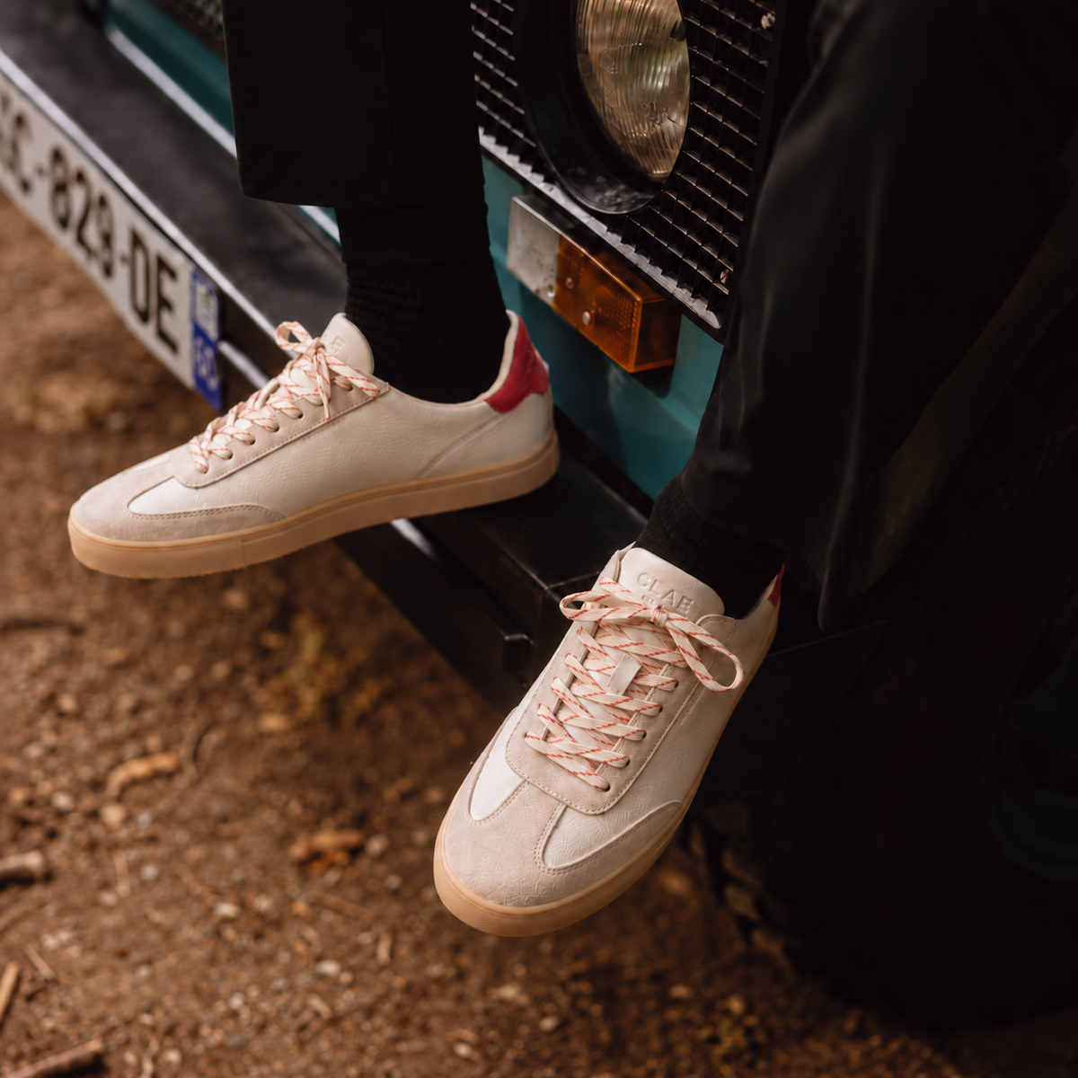 CLAE DEANE Off-White Molten Lava sneakers with red heel tabs and gum soles worn beside a teal vehicle